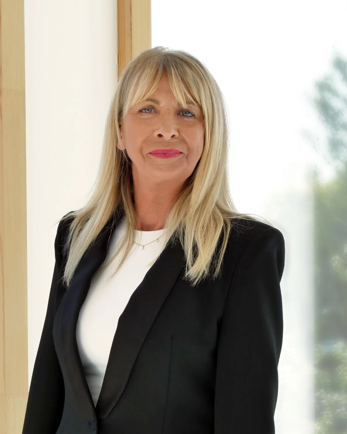 Porträt einer Frau mit blondem Haar im Business-Look vor einem hellen Hintergrund mit Fensterausblick.